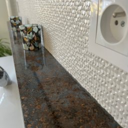 IB-MAX sp. z o.o. - Biała ściana z dekoracyjną mozaiką 3D, brązowy blat z efektem rdzy, dwa świeczniki z motywem kwiatowym i białe gniazdko elektryczne na ścianie.