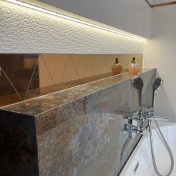 IB-MAX sp. z o.o. - Nowoczesna łazienka z wanną, baterią chromowaną i półką z płytek imitujących rdzę. Nad półką dekoracyjne płytki w odcieniach brązu i bieli z oświetleniem LED.