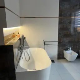 Nowoczesna łazienka z wolnostojącą wanną, bidetem i dekoracyjnymi płytkami imitującymi rdzę, oświetlona półka z kosmetykami.