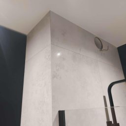 IB-MAX sp. z o.o. - Ściana narożna w łazience wyłożona jasnymi, kwadratowymi płytkami ceramicznymi o delikatnym wzorze, widoczny otwór na oświetlenie z wystającymi przewodami, fragment czarnej ściany i kabiny...