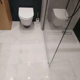 Nowoczesna łazienka z wiszącą toaletą, drewnianą szafką i kabiną prysznicową typu walk-in z odpływem liniowym w podłodze wyłożonej jasnymi, kwadratowymi płytkami.