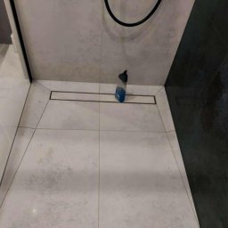 IB-MAX sp. z o.o. - Wrocław: Ułożone jasnoszare płytki podłogowe z odpływem liniowym w kabinie prysznicowej, widoczna butelka płynu do czyszczenia i fragment czarnej płytki na ścianie.