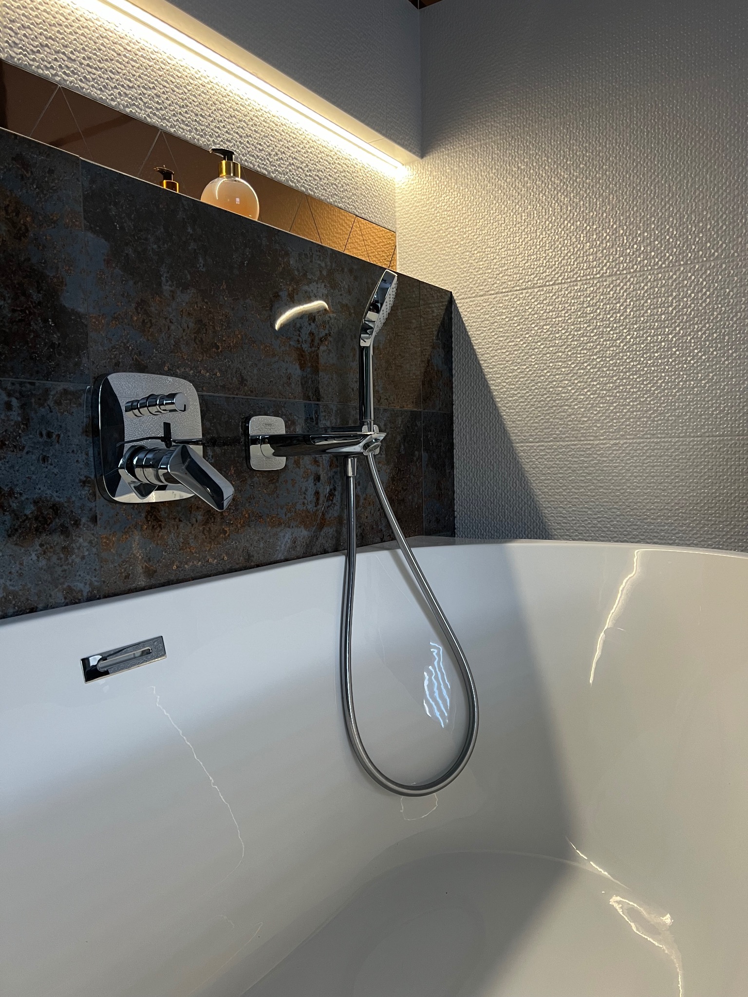Nowoczesna łazienka z wolnostojącą wanną, chromowaną armaturą i ciemnymi płytkami imitującymi kamień, oświetlona listwą LED, z widocznym dozownikiem mydła na półce.