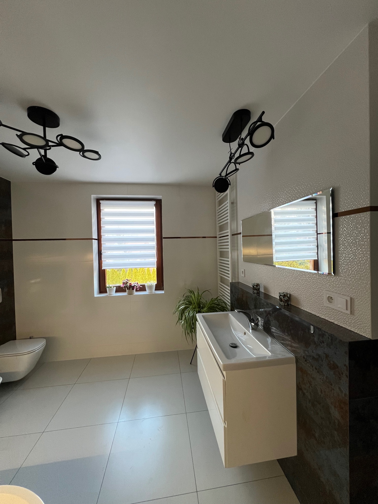 Nowoczesna łazienka z białymi płytkami, wiszącą szafką z umywalką, dużym lustrem, oknem z roletami i designerskim oświetleniem sufitowym w stylu industrialnym.