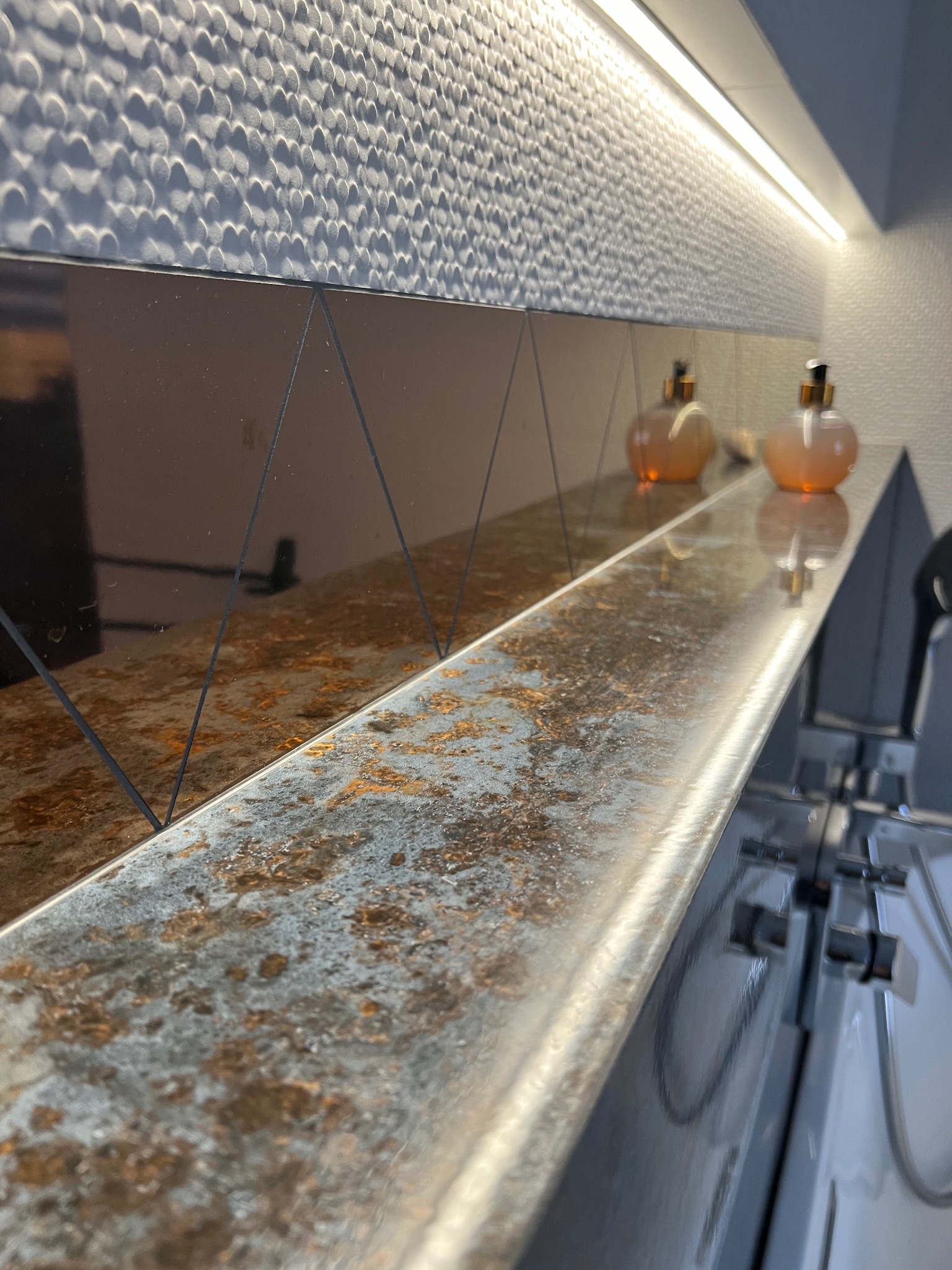 Błyszcząca półka z efektem rdzy, nad nią lustro z geometrycznym wzorem i dekoracyjna ściana z oświetleniem LED, dwie szklane butelki na blacie.