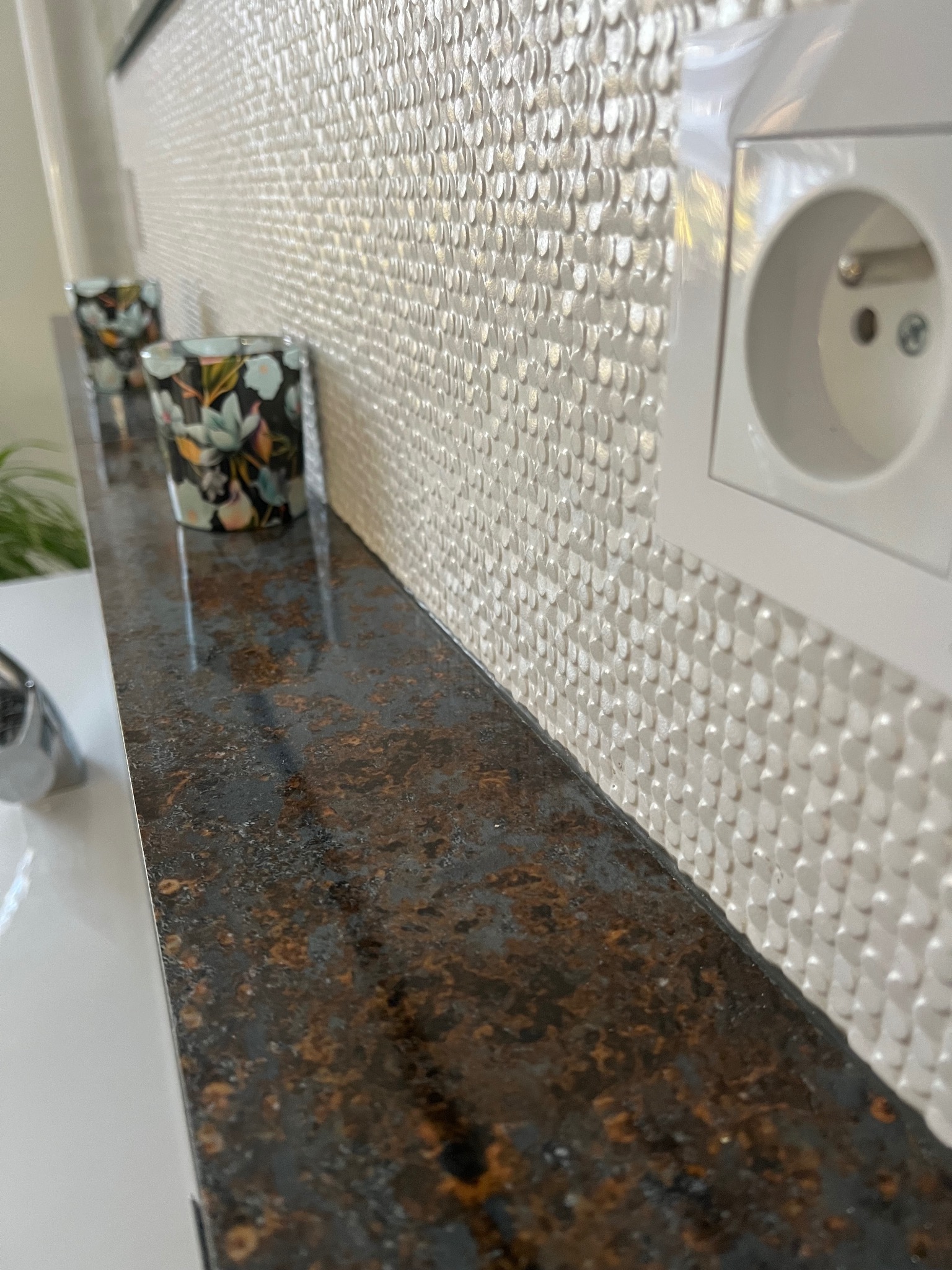 Biała ściana z dekoracyjną mozaiką 3D, brązowy blat z efektem rdzy, dwa świeczniki z motywem kwiatowym i białe gniazdko elektryczne na ścianie.