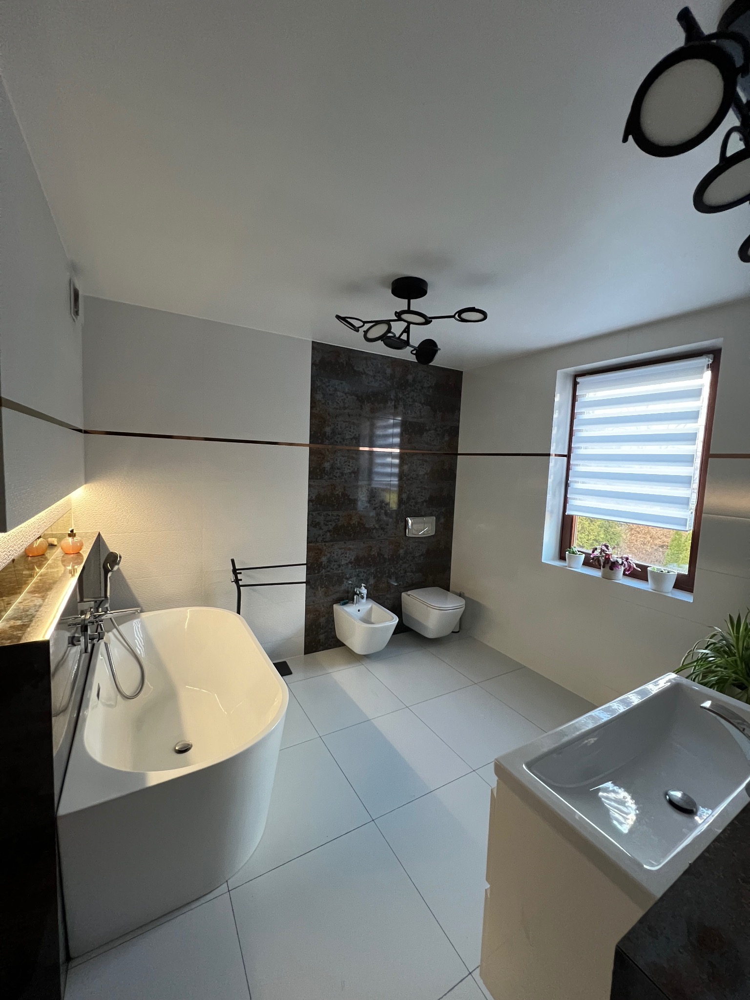 Nowoczesna łazienka z wolnostojącą wanną, bidetem, toaletą i umywalką, wykończona białymi płytkami i ciemną mozaiką, oświetlona designerskimi lampami.