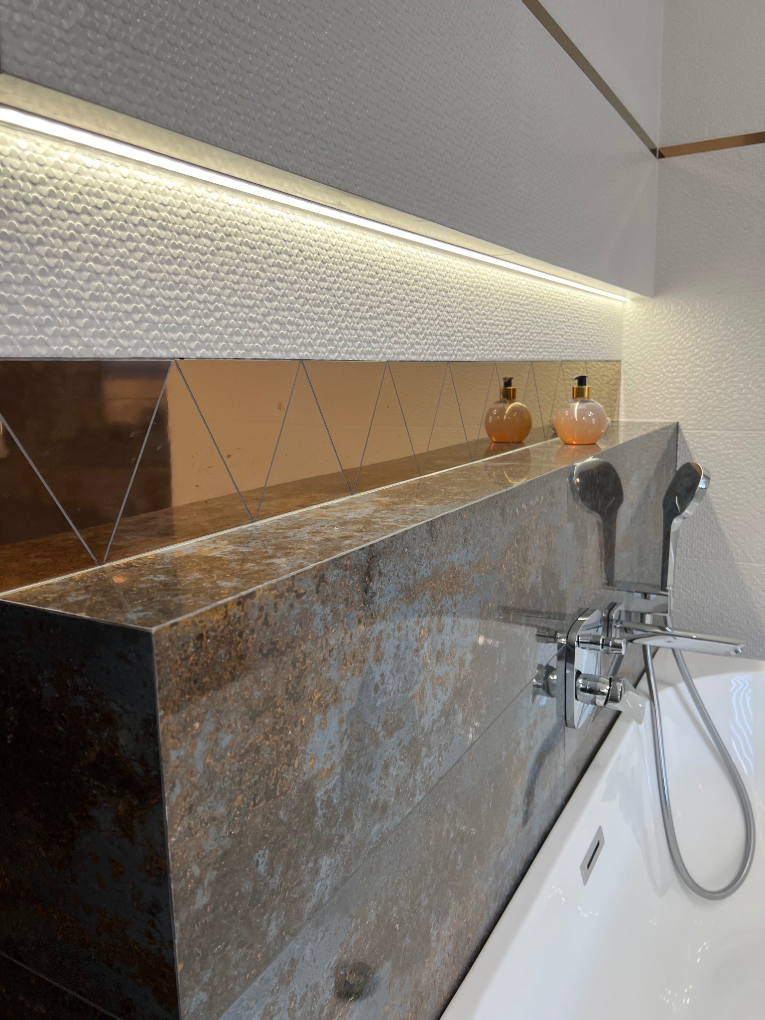Nowoczesna łazienka z wanną, baterią chromowaną i półką z płytek imitujących rdzę. Nad półką dekoracyjne płytki w odcieniach brązu i bieli z oświetleniem LED.