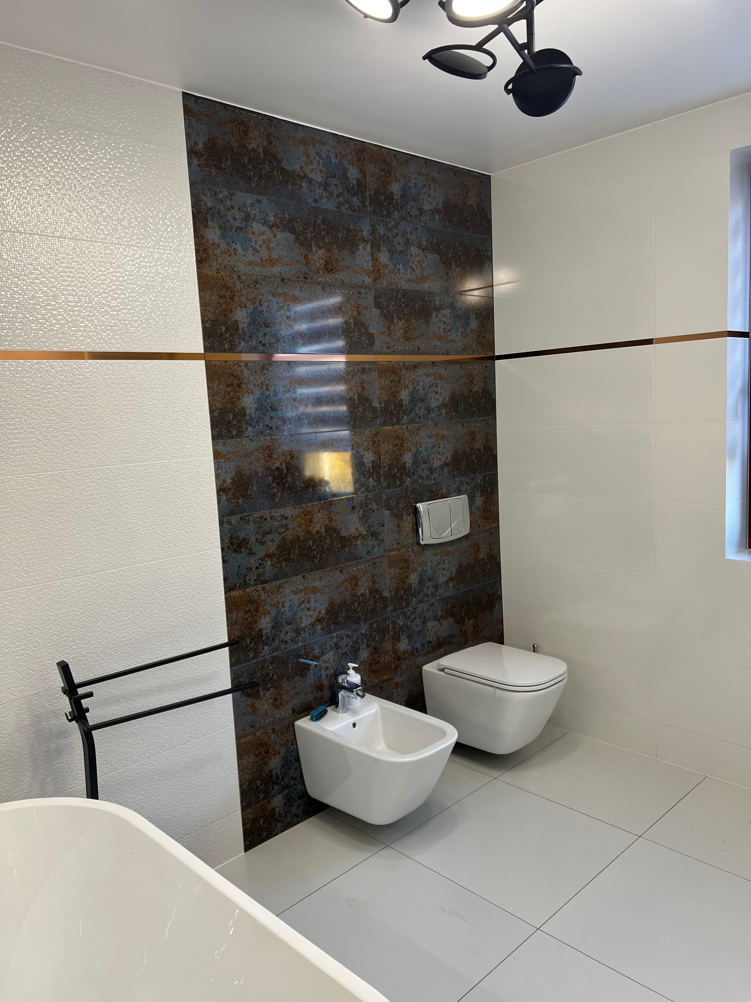 Nowoczesna łazienka z białymi i rdzawymi płytkami, wisząca toaleta i bidet, oświetlenie sufitowe.