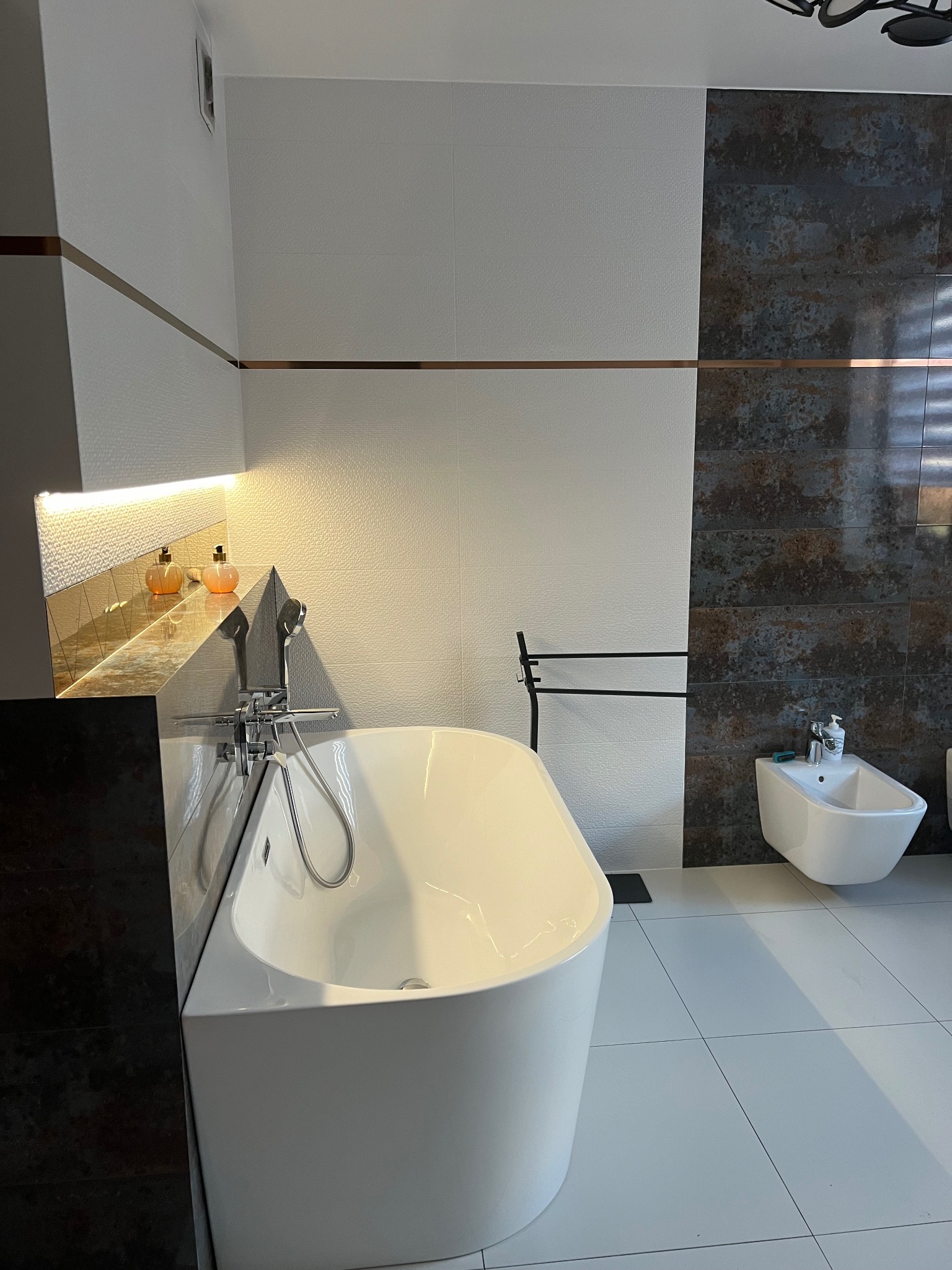 Nowoczesna łazienka z wolnostojącą wanną, bidetem i dekoracyjnymi płytkami imitującymi rdzę, oświetlona półka z kosmetykami.