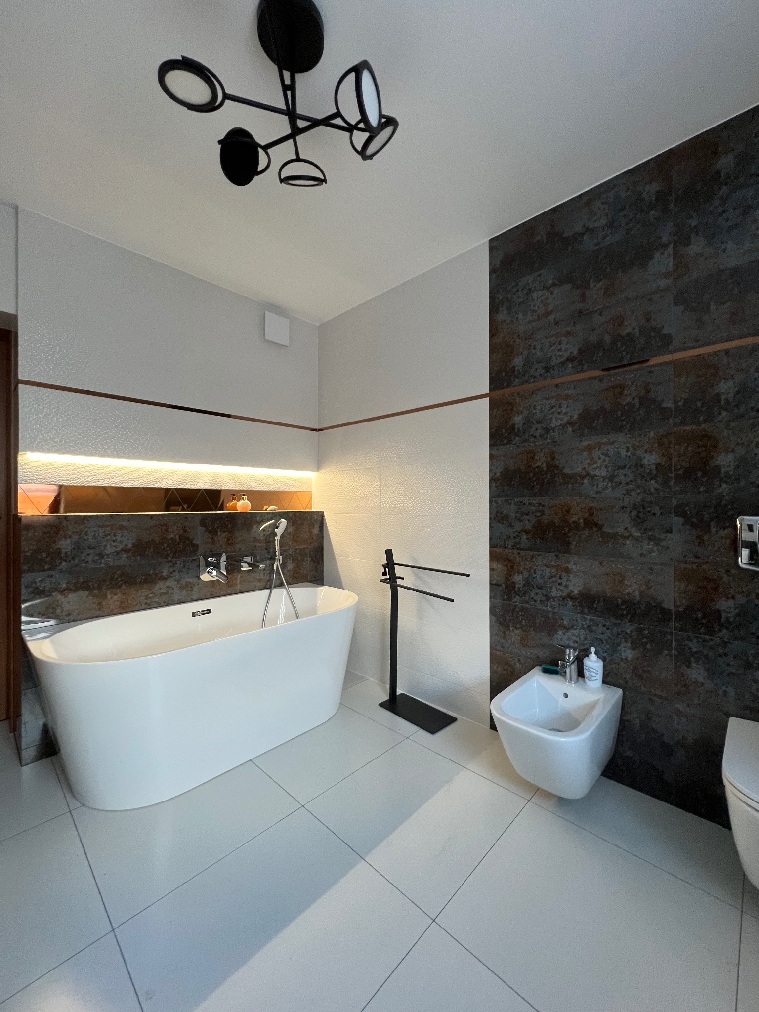 Nowoczesna łazienka z wolnostojącą białą wanną, bidetem i ciemnymi płytkami imitującymi rdzę na ścianie, podświetlona półka z dekoracjami i designerska lampa sufitowa.