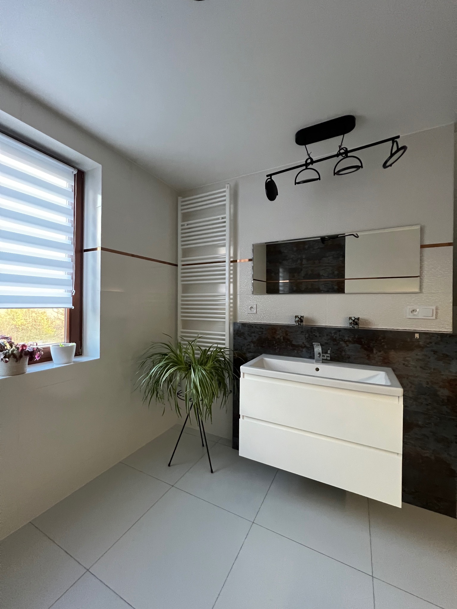 Nowoczesna łazienka z białymi płytkami, umywalką z szafką, lustrem, grzejnikiem drabinkowym i oknem z roletami dzień-noc, oświetlona czarną lampą sufitową i ozdobiona roślinami doniczkowymi.