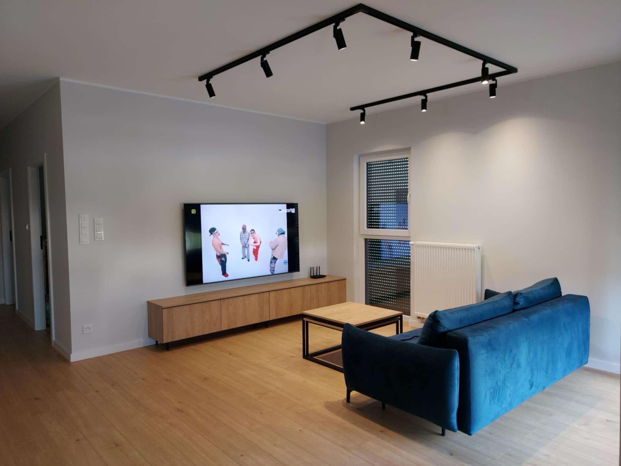 Salon z drewnianą podłogą, szarą ścianą, telewizorem, szafką RTV i niebieską sofą, oświetlony reflektorami na szynoprzewodzie.