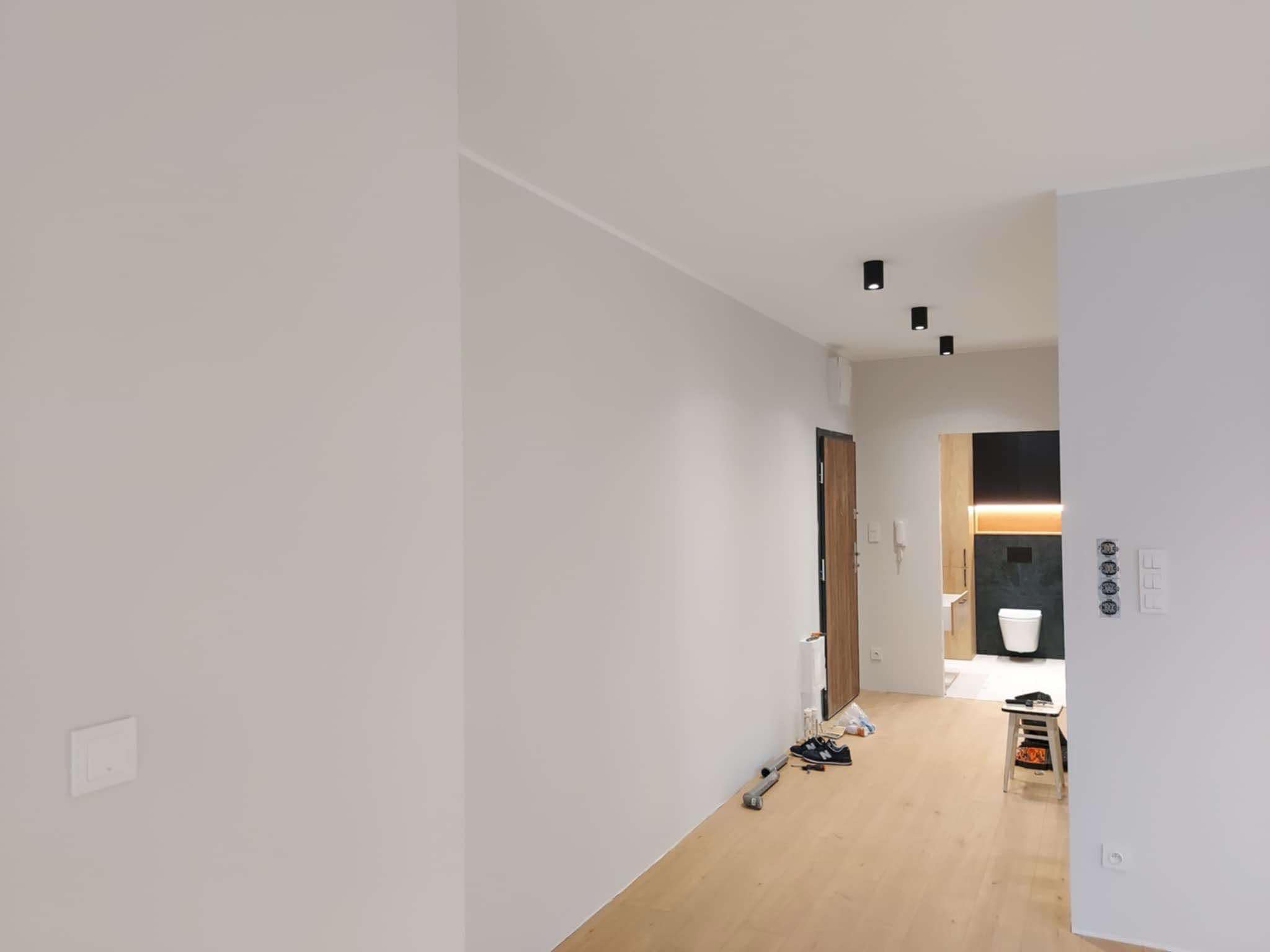 Wnętrze mieszkania po remoncie: Jasne, pomalowane ściany w odcieniach bieli i szarości, drewniana podłoga, czarne lampy sufitowe, widok na łazienkę z toaletą i czarną ścianą, widoczne gniazdka...