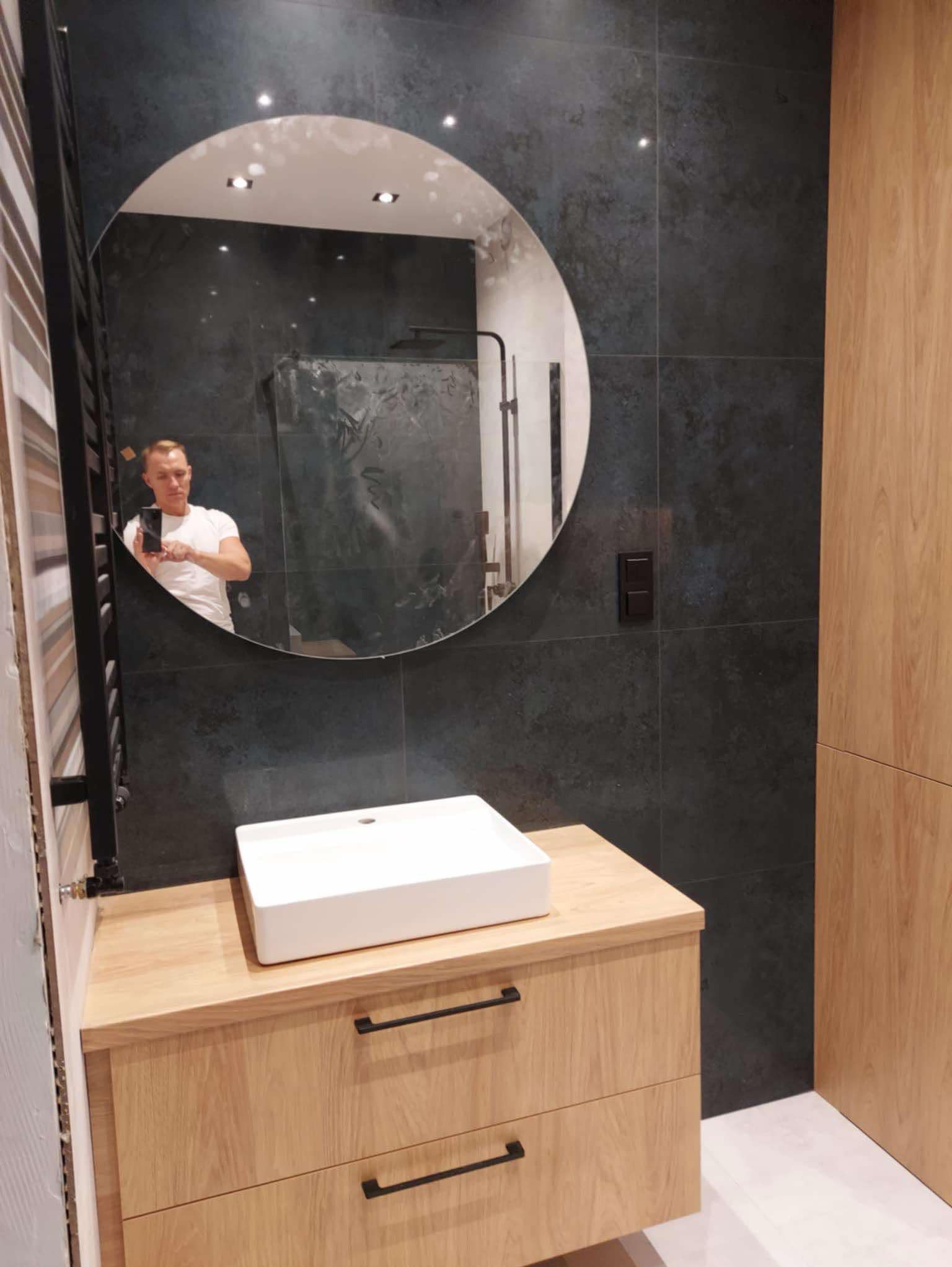 Nowoczesna łazienka z ciemnymi płytkami, okrągłym lustrem, białą umywalką nablatową na drewnianej szafce z czarnymi uchwytami; odbicie fotografa w lustrze.