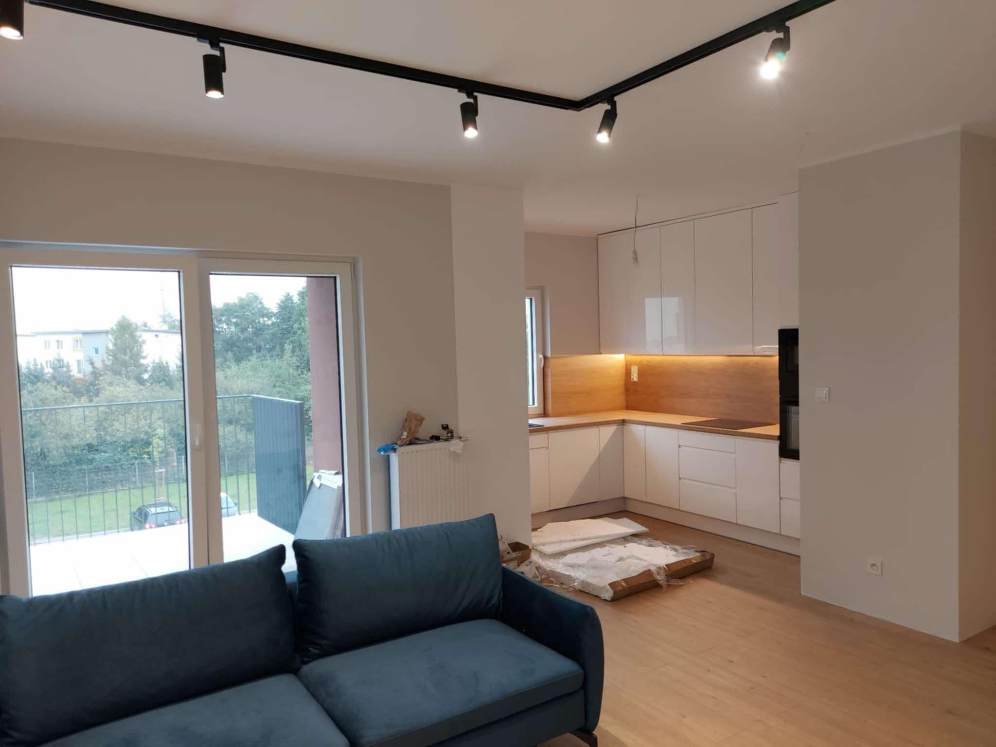 Salon z aneksem kuchennym w trakcie wykańczania, widoczna niebieska sofa, białe szafki kuchenne z drewnianym blatem, oświetlenie szynowe na suficie i częściowo rozpakowane płyty na podłodze.