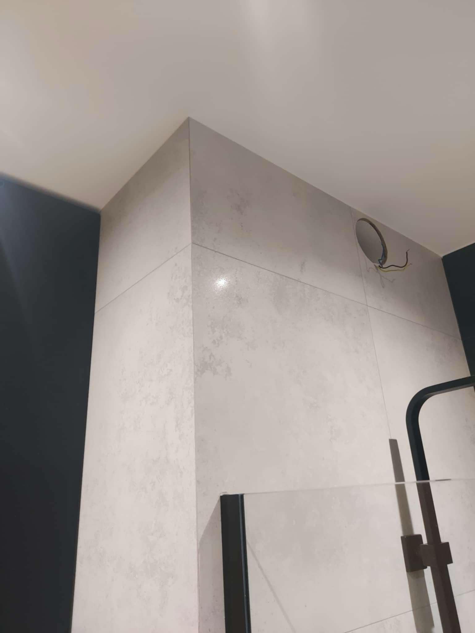 Ściana narożna w łazience wyłożona jasnymi, kwadratowymi płytkami ceramicznymi o delikatnym wzorze, widoczny otwór na oświetlenie z wystającymi przewodami, fragment czarnej ściany i kabiny...