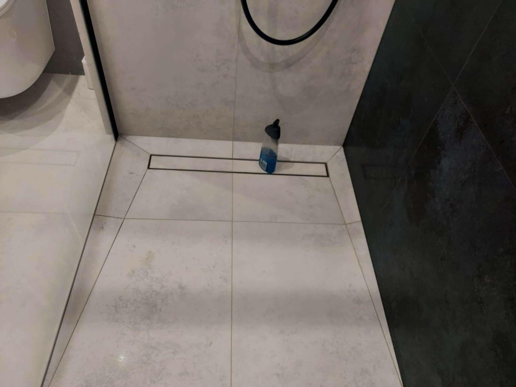 Wrocław: Ułożone jasnoszare płytki podłogowe z odpływem liniowym w kabinie prysznicowej, widoczna butelka płynu do czyszczenia i fragment czarnej płytki na ścianie.