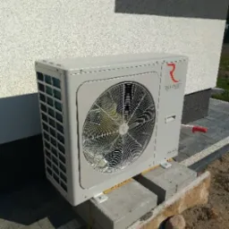 Zewnętrzna jednostka klimatyzacji inwerterowej zamontowana na betonowych bloczkach przy ścianie budynku z jasnym tynkiem.