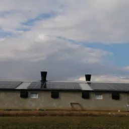 Budynek gospodarczy z panelami fotowoltaicznymi na dachu, kominami wentylacyjnymi i dwoma srebrnymi silosami w tle, częściowo pochmurne niebo.