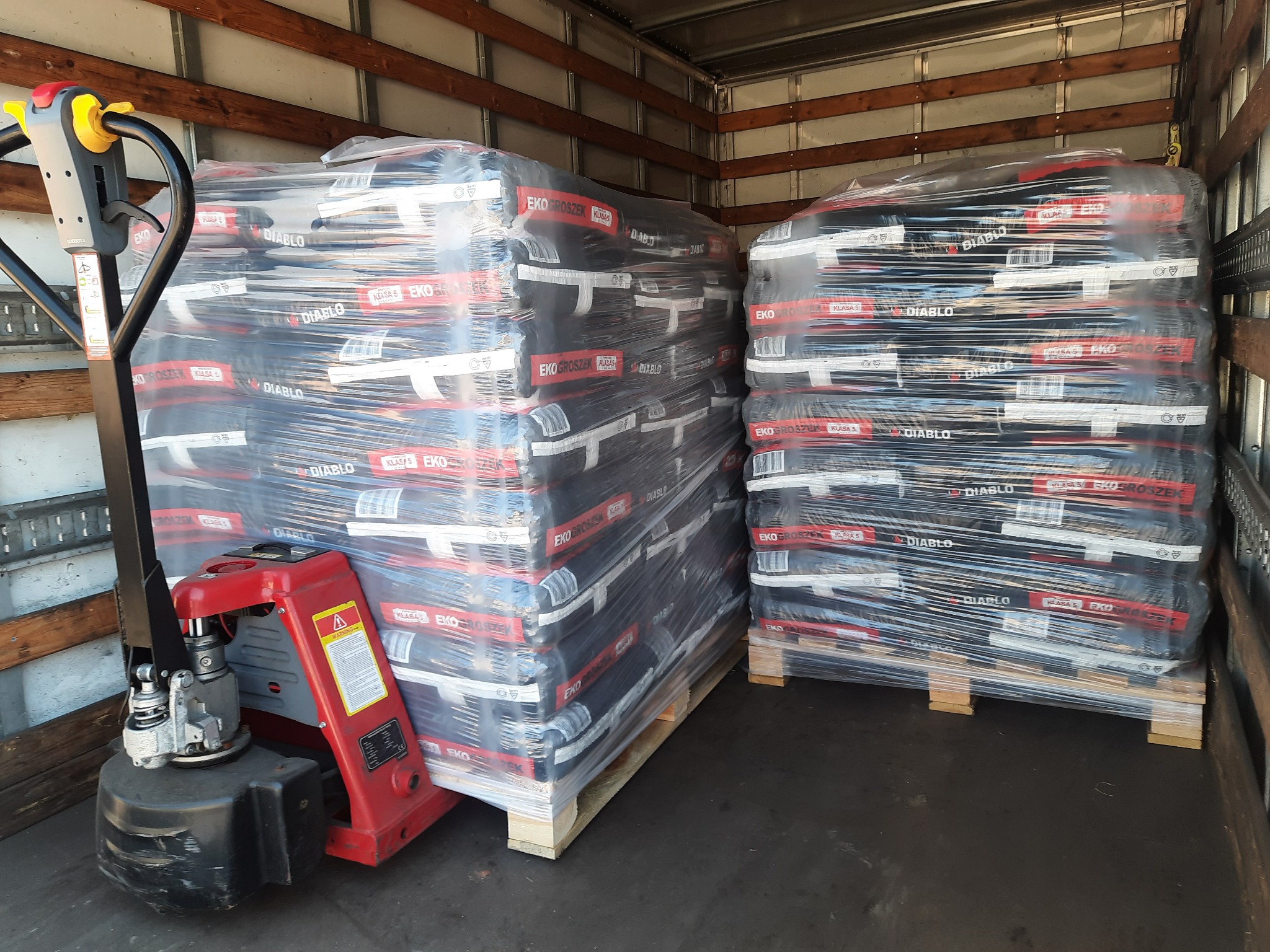 Wnętrze naczepy z paletami worków z ekogroszkiem, zabezpieczonych folią stretch, z widocznym wózkiem paletowym.