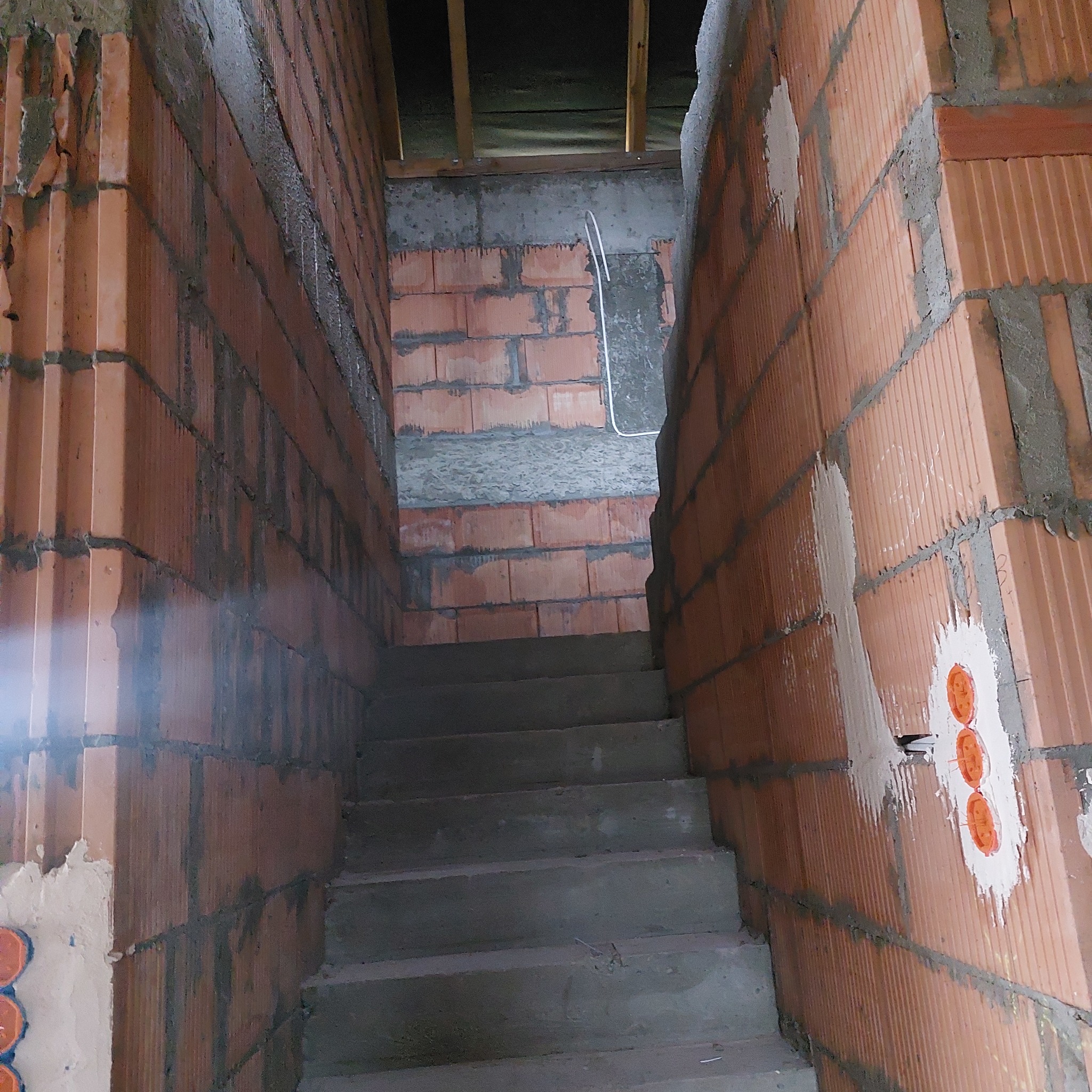 Murowane schody w surowym stanie, ściany z czerwonej cegły z widocznymi przewodami elektrycznymi i niedokończonymi tynkami.