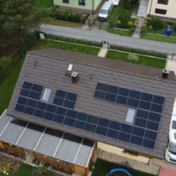 Widok z drona na dom jednorodzinny z brązową dachówką, na której zamontowane są panele słoneczne, obok domu zaparkowane samochody i kamper.