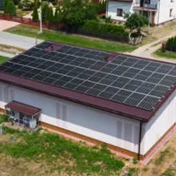 Widok z góry na budynek z panelami słonecznymi na dachu z brązowej blachy, otoczony zielenią i innymi budynkami.