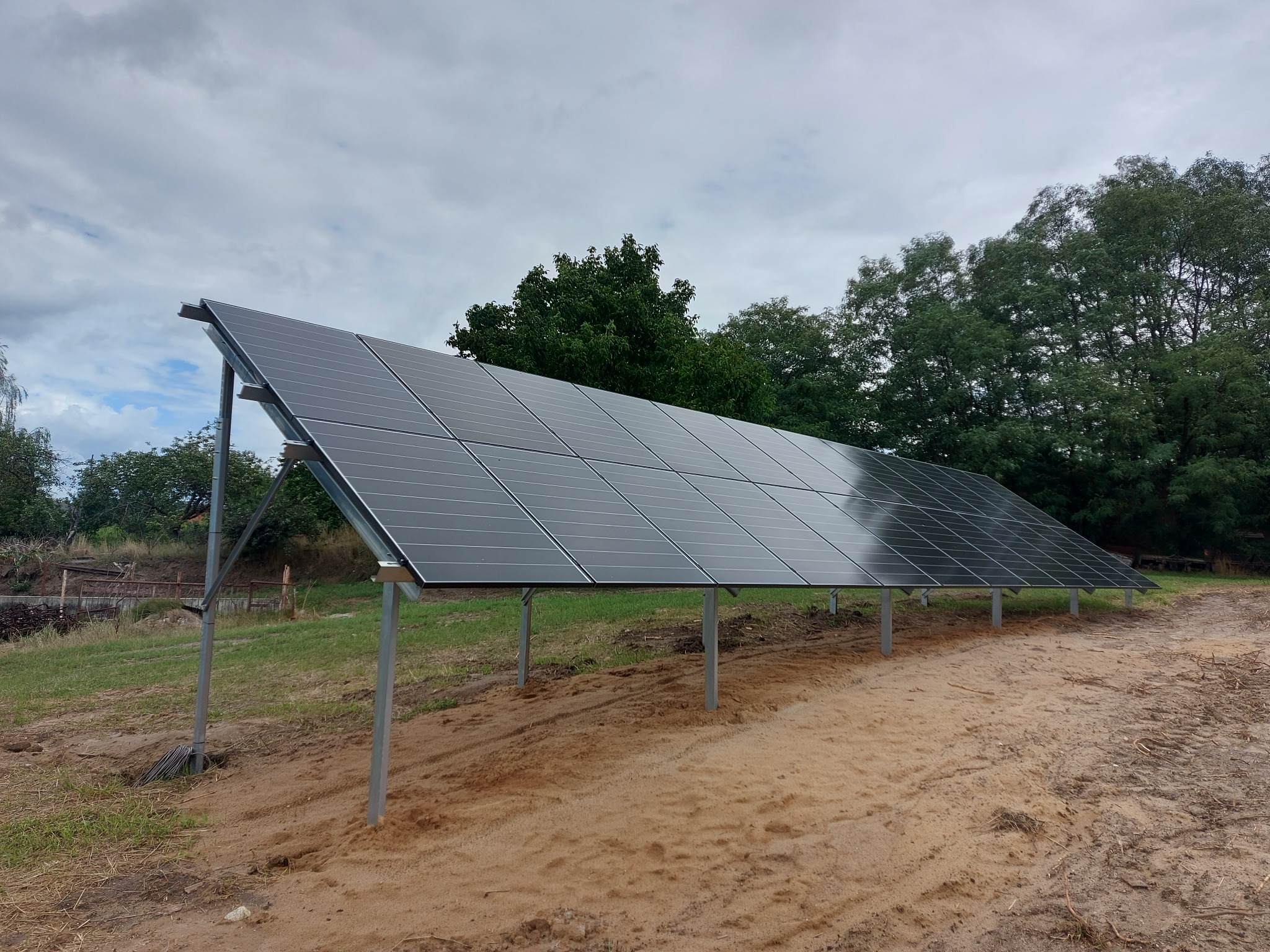 Rząd paneli słonecznych na metalowych podporach, zainstalowanych na piaszczystym gruncie, z zielenią drzew w tle pod pochmurnym niebem.