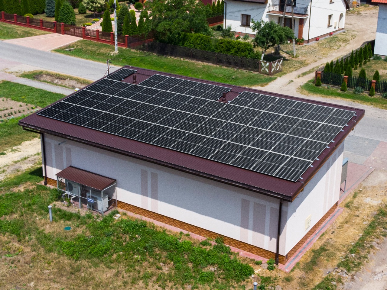 Widok z góry na budynek z panelami słonecznymi na dachu z brązowej blachy, otoczony zielenią i innymi budynkami.