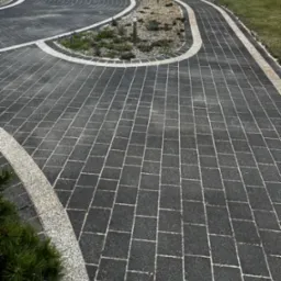 Czarna kostka brukowa ułożona w geometryczny wzór, obrzeża z jasnego kamienia, rabaty z roślinami i oświetleniem w tle, widok na podjazd i ogrodzenie.