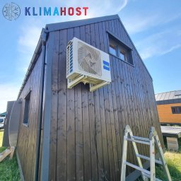 Klimahost - Zewnętrzna jednostka klimatyzacji Haier zamontowana na ciemnej, drewnianej elewacji domu modułowego, widoczna drabina aluminiowa i logo firmy Klimahost w górnym rogu.
