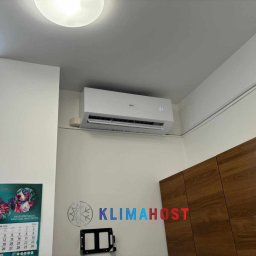 Klimahost - Biały klimatyzator Haier zamontowany na ścianie w pomieszczeniu, widoczny wyświetlacz temperatury, obok szafka z drewnianymi frontami i kalendarz z motywem psów.