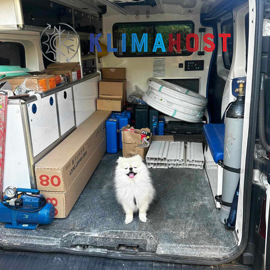 Wnętrze samochodu serwisowego wypełnione materiałami do instalacji klimatyzacji, takimi jak rury, skrzynki, butle z gazem i pompa próżniowa; w centrum kadru biały pies rasy szpic.