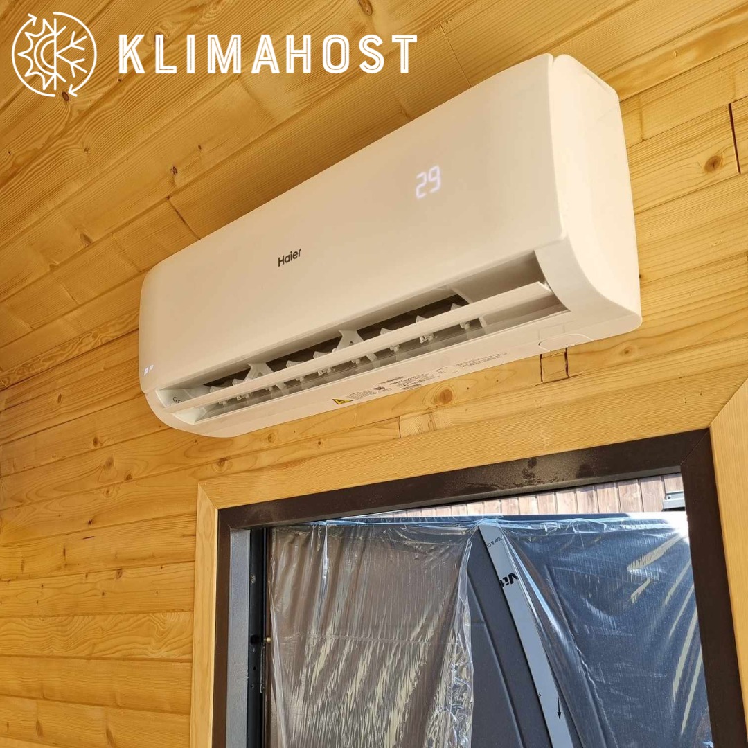 Biały klimatyzator Haier zamontowany na ścianie z drewnianych paneli, z wyświetlaczem wskazującym temperaturę 29 stopni, widoczne logo firmy Klimahost w lewym górnym rogu, okno w folii ochronnej...