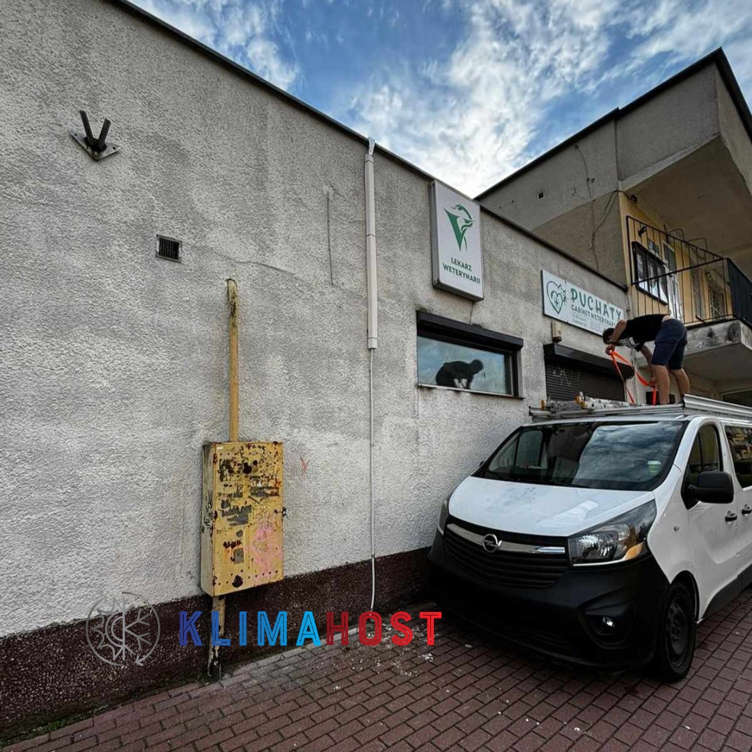 Pracownik na drabinie na dachu białego samochodu serwisowego, montujący lub serwisujący urządzenie na elewacji budynku z szyldem gabinetu weterynaryjnego.