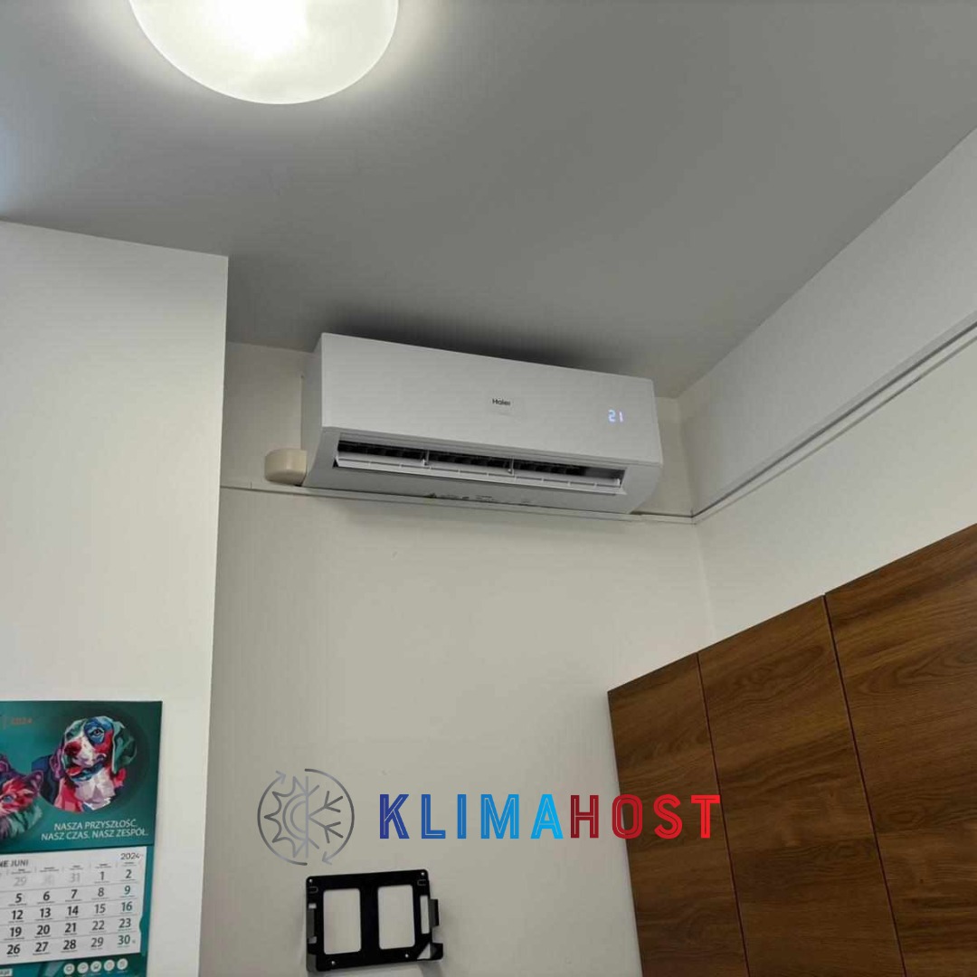 Biały klimatyzator Haier zamontowany na ścianie w pomieszczeniu, widoczny wyświetlacz temperatury, obok szafka z drewnianymi frontami i kalendarz z motywem psów.
