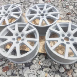 Cztery aluminiowe felgi samochodowe po piaskowaniu i malowaniu proszkowym na szaro, ułożone na drewnianej płycie na tle gruzu.