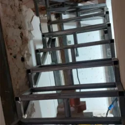 Metalowa konstrukcja schodów w surowym stanie, spawana, z widocznym sprzętem spawalniczym, w trakcie montażu w budynku z odsłoniętą cegłą i fragmentami tynku.