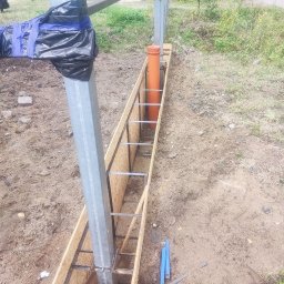 Yarmar Marta Fedoriv - Metalowe słupy w drewnianej szalunku, przygotowane do zalania betonem. Widoczna pomarańczowa rura. Prace ziemne w toku, teren trawiasty w tle.