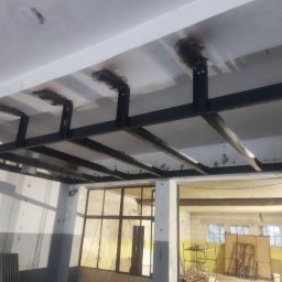 Yarmar Marta Fedoriv - Metalowa konstrukcja podwieszana do sufitu, spawana, pomalowana na czarno. Widoczne spawy i mocowania do sufitu. W tle okna i elementy wyposażenia warsztatu.