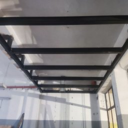 Yarmar Marta Fedoriv - Czarna, metalowa konstrukcja sufitowa z profili, widoczna z dołu, w jasnym pomieszczeniu. Elementy spawane, surowy wygląd, widoczne ślady montażu.