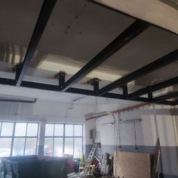 Yarmar Marta Fedoriv - Metalowa konstrukcja podwieszana pod sufitem w hali, widoczne spawane elementy i mocowania. W tle okna i wyposażenie warsztatu.