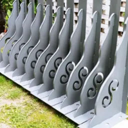 Szereg pomalowanych na szaro, metalowych elementów balustrady z wyciętym wzorem, ustawionych na trawie przed ceglanym murem.