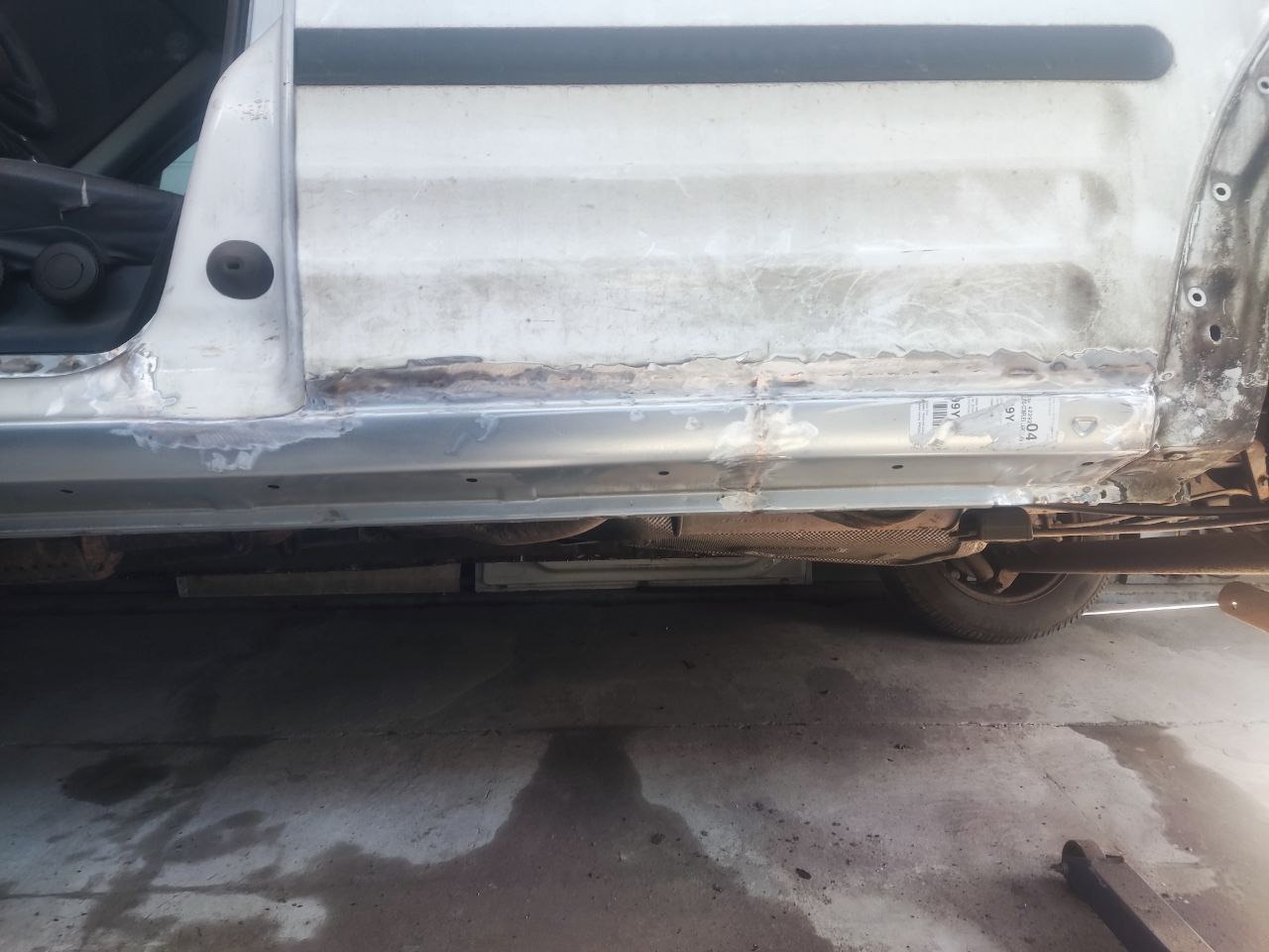 Naprawa progu białego samochodu w warsztacie. Widoczne ślady spawania i obróbki blacharskiej. Dolna część karoserii po naprawie, z widocznymi elementami podwozia.