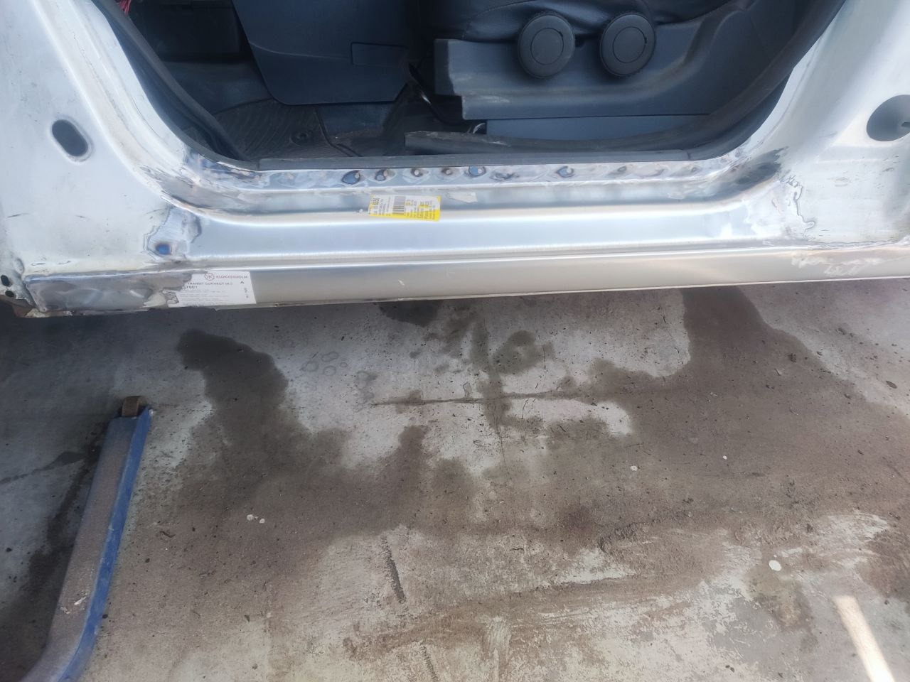 Naprawa progu samochodu: widoczne ślady spawania i szlifowania. Próg w kolorze srebrnym, z widocznymi naklejkami informacyjnymi. Podłoga warsztatu zabrudzona.