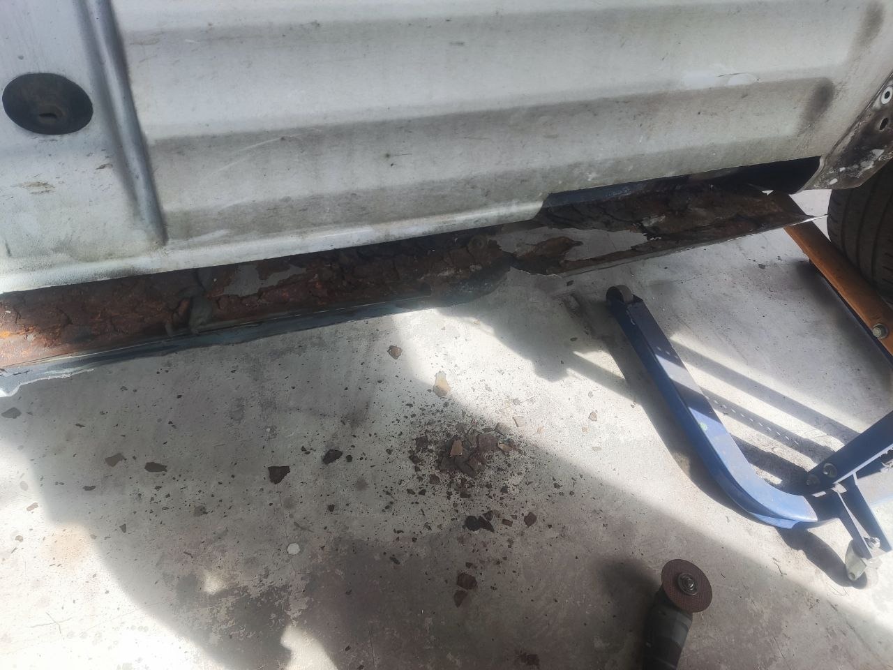 Korozja progu białego samochodu w warsztacie, widoczny podnośnik i szlifierka kątowa, fragment opony. Rdza na metalu.