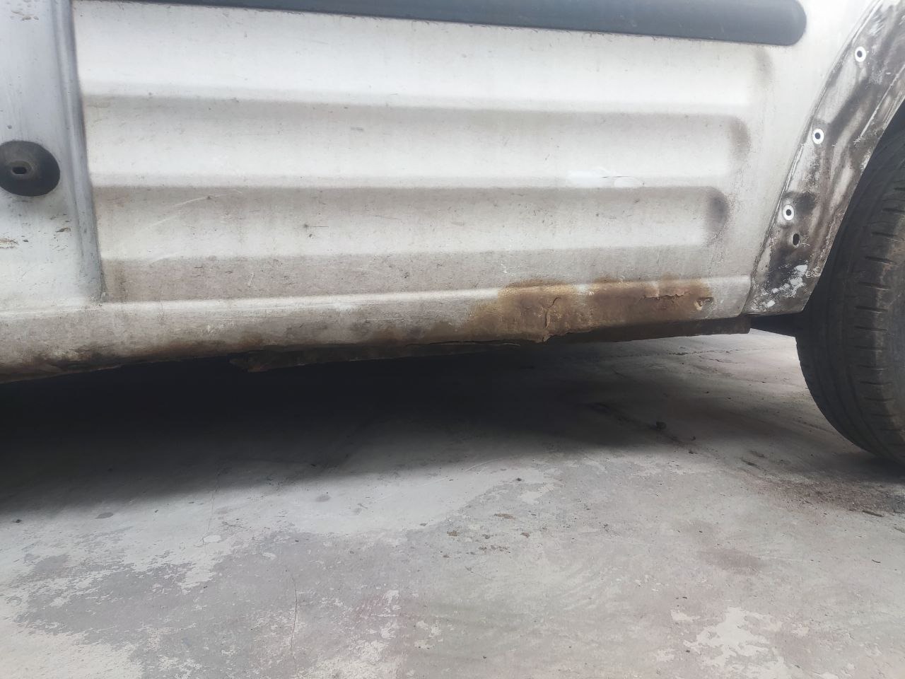 Bok samochodu z widoczną korozją na progu i nadkolu. Przygotowanie do naprawy, widoczne otwory po usuniętych elementach.