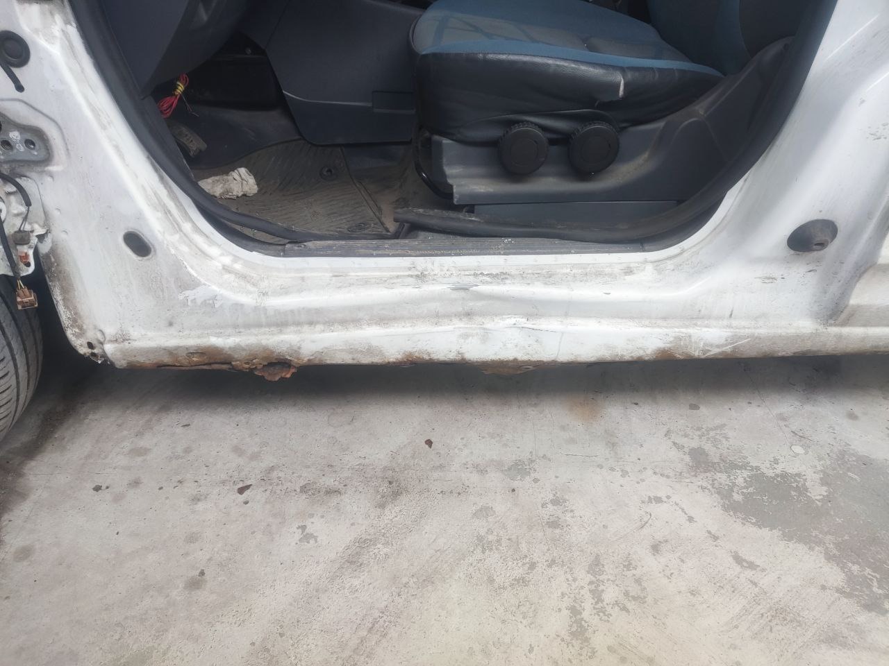 Biały samochód z widoczną korozją na progu, wnętrze kabiny z niebieskim fotelem. Ukazany stan przed naprawą blacharską, zarys rdzy na dolnej części karoserii.