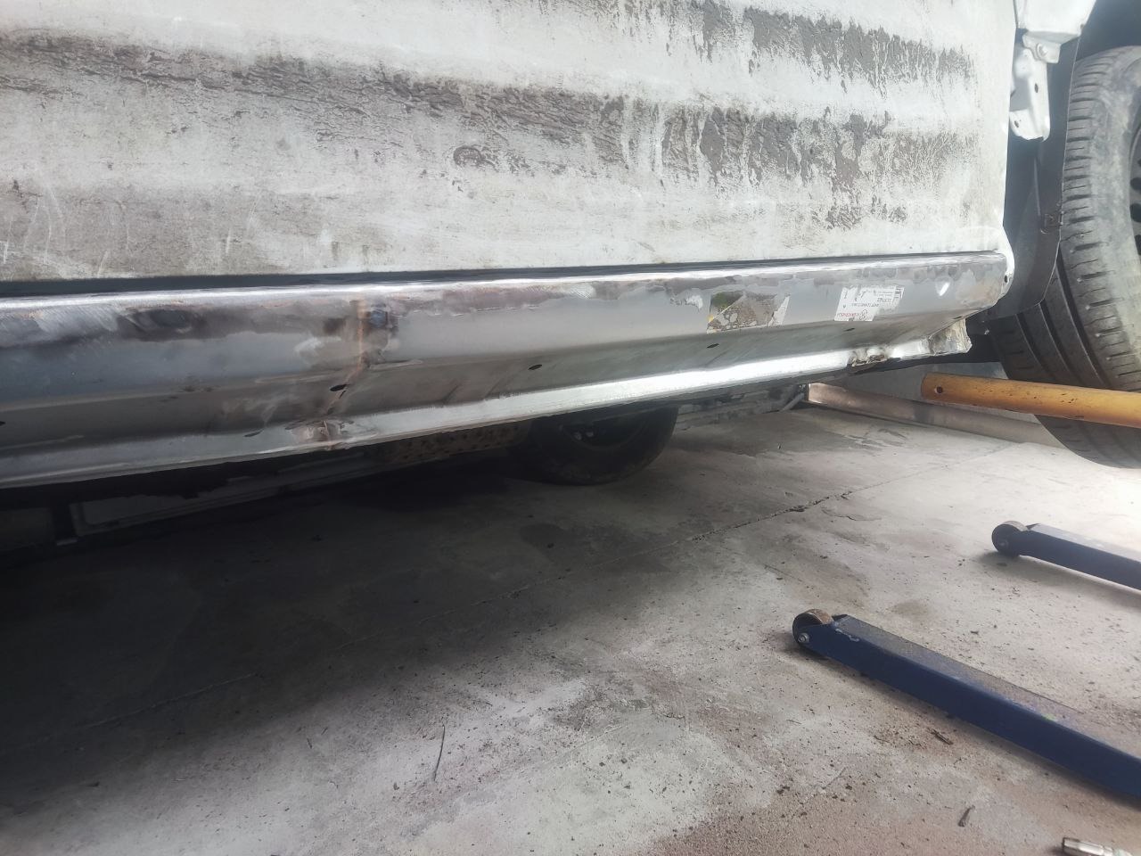 Fragment karoserii samochodu w trakcie naprawy, widoczne ślady szpachlowania i spawania. Podnośniki i opona w tle. Blacha przygotowana do lakierowania.